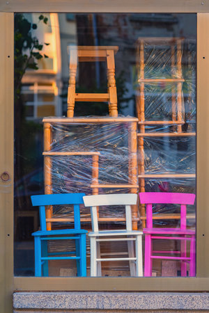 Colorful wooden children chairs stand in shop window display, with natural wooden stools stacked and wrapped in plastic behind them. Concept of furniture, design, chair variety, urban retail displayの写真素材