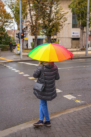 Woman with colorful rainbow umbrella standing at city crosswalk on rainy autumn day. Concept of rainy weather, urban lifestyle, autumn moodの写真素材
