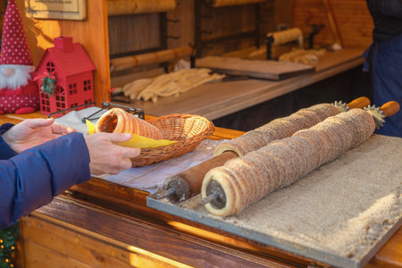 Hands of buyer with traditional chimney cake prepared fresh at street food market, with sugar-coated dough rolled on wooden spindles. Concept of sweet pastry, festive treat, authentic food experienceの写真素材