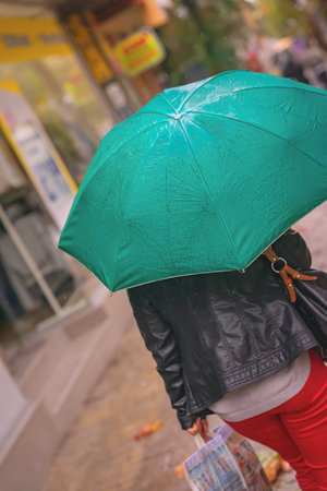 Woman rushing with green teal umbrella on rainy urban street. Intentional rotation of angle towards horizon. Concept of autumn weather, lifestyle, city rain mood, street life in motionの写真素材