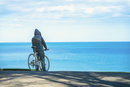 Back view of person in hoodie with bicycle looking at calm blue sea under a bright cloudy sky. Concept of solitude, freedom of movement, natureの写真素材