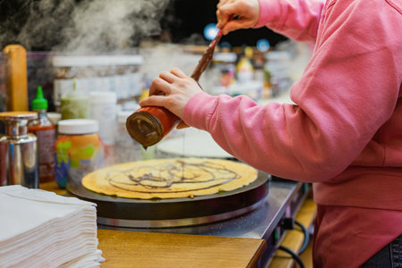 Chocolate crepe preparation at market. Person in pink hoodie preparing hot crepe with chocolate sauce, festive food stall. Concept of street food, sweet cravings, cozy winter treats, Christmas marketの写真素材