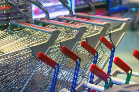 Shopping Carts in Store. Row of metal shopping carts with red handles stacked inside bright supermarket. Concept of consumerism, retail logistics, shopping routineの写真素材