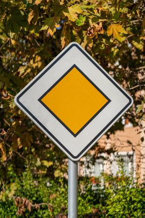 Priority road sign in front of autumn foliage. Yellow diamond-shaped priority road sign stands out against background of autumn trees and sunny day. Concept of traffic regulation, road safety signalの写真素材