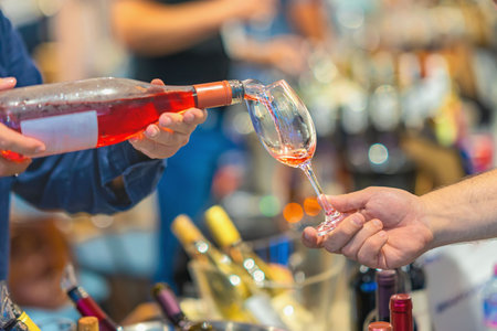 Close-up of hands with Rose Wine poured into wine glass, tasting event, bottles of wine in...