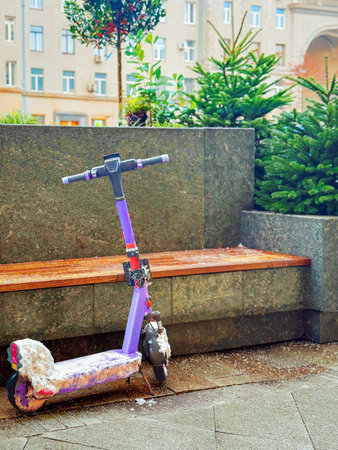 Electric scooter covered with snow parked near bench in city square. Concept of urban mobility, seasons, winter transportの写真素材