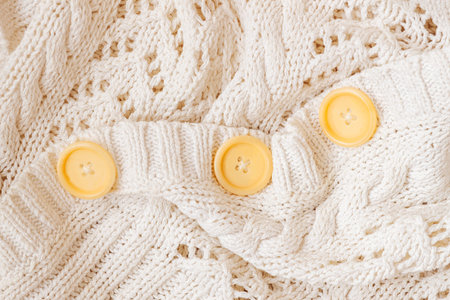 White knitted pullover fabric with cable knit pattern, round buttons close-up, clothing detail,...