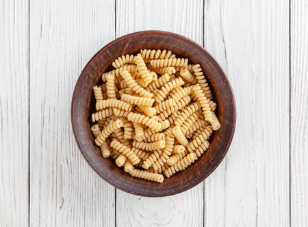 Uncooked fusilli pasta in ceramic bowl on white wooden backgroundの写真素材