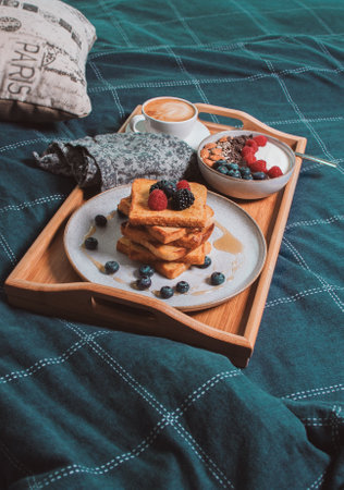 Cozy breakfast in bed, yogurt bowl with berries and nuts, french toasts, coffee cup on wooden plate