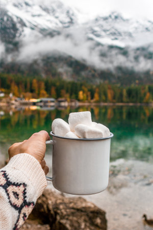 Picnic in the Bavarian mountains, Germany. Hand with mug of cacao and marshmallow on the lake, forest backgroundの写真素材