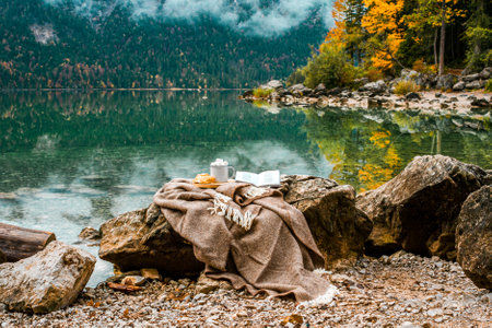 Picnic on stones in the Bavarian mountains, Germany. Plaid, cacao with marshmallow, croissant, book near lake and autumn forest.の写真素材