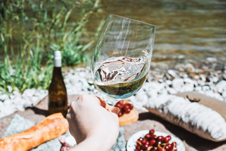 Hand with white wine glass. Romantic rustic picnic on the beach, bottle, baguette, cherries, grapeの写真素材