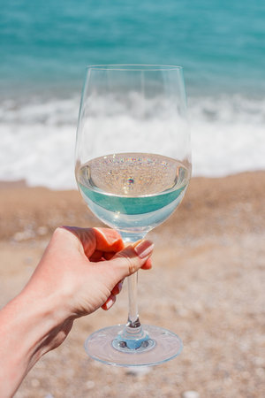 Woman hand with white wine glass on a turquoise Mediterranean sea foam, waves and beach backgroundの写真素材