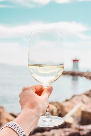 Woman hand with white wine glass on a sea, stones and lighthouse background. Nice, French rivieraの写真素材