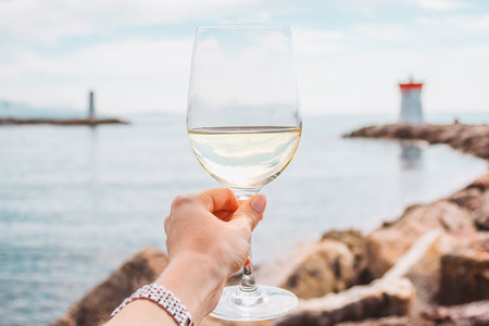Woman hand with white wine glass on a sea, stones and lighthouse background. Nice, French rivieraの写真素材