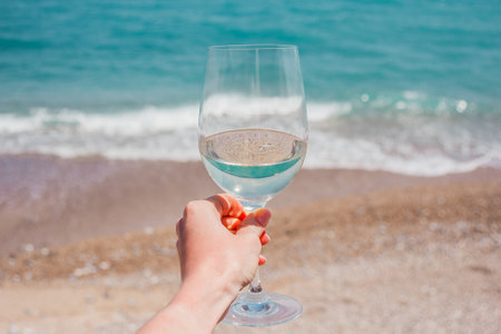 Woman hand with white wine glass on a turquoise Mediterranean sea foam, waves and beach backgroundの写真素材