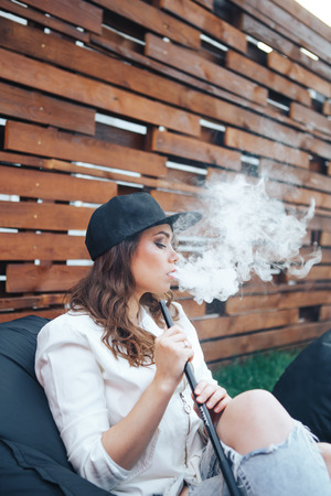 Young woman in a black cap smoking a hookah outdoors. The pleasure of smoking. Background a wooden wall.の写真素材