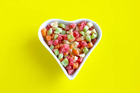 Candied fruits multicolored on a platter in the shape of a heart on a yellow background top view.の写真素材