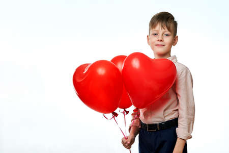 European boy holds three inflatable heart-shaped balloons, a Valentine's Day gift, dressed in a shirt and trousers, against a white insulated backgroundの写真素材