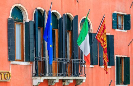Facade of a typical old building with shutters and ornate windows, the Italian, the European and the Venitian flags in Venice, Italyの写真素材