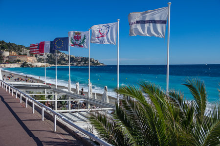 Nice, France - October 11, 2013: View of bright blue water and Mediterranean Sea coastline with various flags along the Promenade des Anglais & a palm tree in the foreground, Nice, Cote d'Azur, Franceのeditorial素材