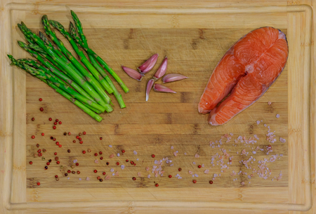 Fresh salmon steak surrounded by pink peppercorns, Himalayan salt, garlic and asparagus on a wooden cutting boardの写真素材