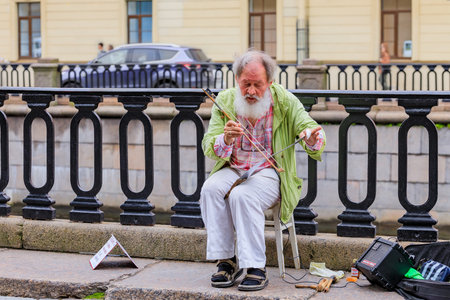 Saint Petersburg, Russia - September 10, 2017: Old male street performer with a beard, dressed in shabby clothing playing a hand saw for money on the streets of Saint Petersburgのeditorial素材