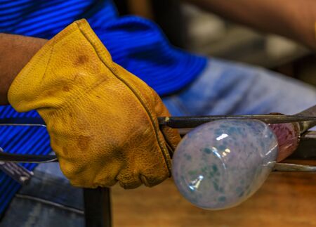 Glass blower shaping a bubble of melted glass on a rod by hand in the manufacturing process at a glass maker's workshop, shallow depth of fieldの写真素材