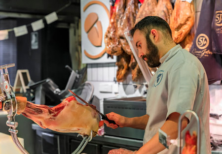 Madrid, Spain - June 4, 2017: Butcher slicing a whole leg of serrano iberico ham on a stand for customers at a market, hams hanging in the backgroundのeditorial素材