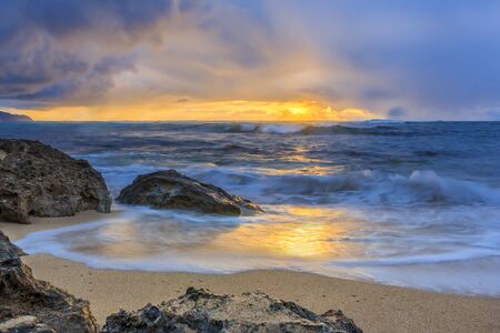Waves rolling onto a rocky beach with a stunning sunset and rain in the background in North Shore,...