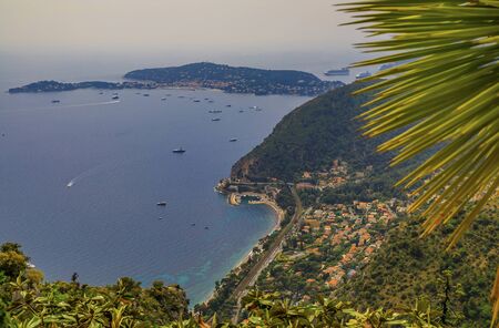 Scenic view of Mediterranean coastline, the Alpes and medieval houses from Eze village town on the French Riviera, France with a blurred palm frondの写真素材