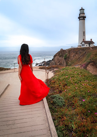 Yong beautiful brunette woman in a long elegant red dress posing near the Pacific Ocean on California Coast with a lighthouse in the backgroundの写真素材