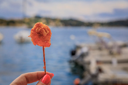 Crispy shrimp fritter on a stick with Mediterranean Sea and boats in background, Villefranche sur Mer Old Town on the French Riviera, South of Franceの写真素材
