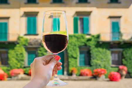 Woman s hand holding a glass of red Chianti wine at a vineyard with an ivy covered stone house at a vineyard in the famous Tuscany region, Italyの写真素材