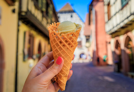 Woman's hand holding artisanal pistachio gelato in a cone, with half timbered in Kaysersberg, a...