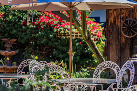 Empty rustic outdoor restaurant table with wrought iron chairs under an umbrella on a patio with blooming red flowers, San Juan Capistrano, Californiaの写真素材