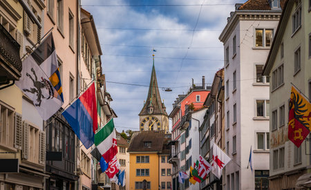 Zurich, Switzerland - April 10, 2024: St. Peter Church clock tower from Rennweg, cobblestone street with colorful guild flags on historic buildingsの写真素材