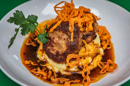 A plated gourmet dish of a meat patty on a bed of mashed potatoes with crispy fried onions and a...