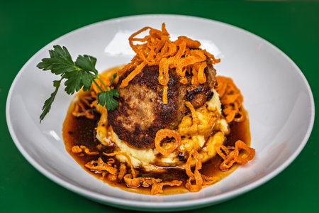A plated gourmet dish of a meat patty on a bed of mashed potatoes with crispy fried onions and a...