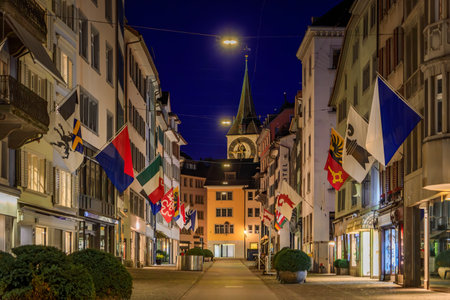 St. Peter Church clock tower from Rennweg, cobblestone street with colorful guild flags hanging on historic buildings at sunset in Zurich, Switzerlandの写真素材