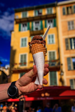 Man s hand holds a cone of artisanal chocolate pistachio gelato, view of traditional colorful...
