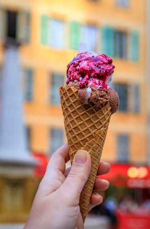 Hand holds a cone of artisanal chocolate and amarena cherry gelato, view of traditional colorful...