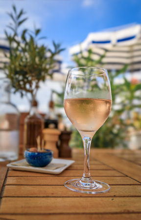 A glass of rose wine at an outdoor waterfront restaurant with a background of the beach umbrellas, blurred olive trees and blue skies in Nice, Franceの写真素材