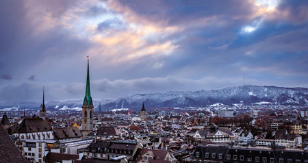 Aerial winter cityscape with iconic church towers, terracotta tile roofs, and snow covered Uetliberg mountain after a snowfall in Zurich, Switzerlandの写真素材