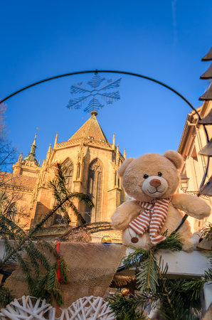 A teddy bear sits in front of a decorative archway at the Christmas Market with the spire of the Colmar Cathedral visible in the background, Franceの写真素材
