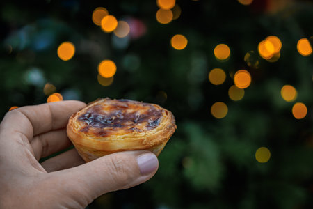 Woman s hand holds golden brown pastel de nata, Portuguese egg custard tart pastry, festive lights...