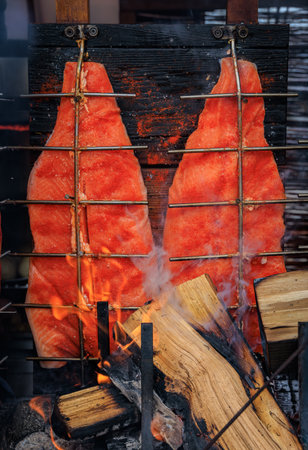 Salmon fillet roasting on wooden planks over open fire, glowing embers and smoke create a rustic...