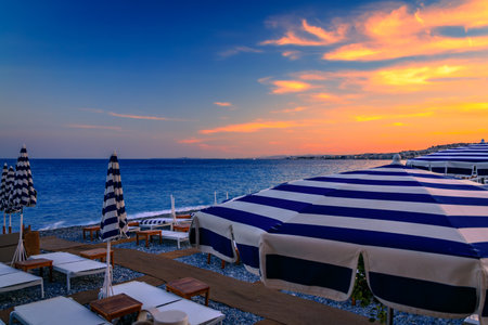 Blue and white striped umbrellas line a pebbled beach during a golden sunset over the Mediterranean Sea along the Promenade des Anglais, Nice, Franceの写真素材