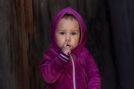 A little girl holds a finger near her lips and shows a gesture of silence. A small child with a worried face holds a finger on his lips, shows a sign of silence, isolated on a dark wooden background.の写真素材