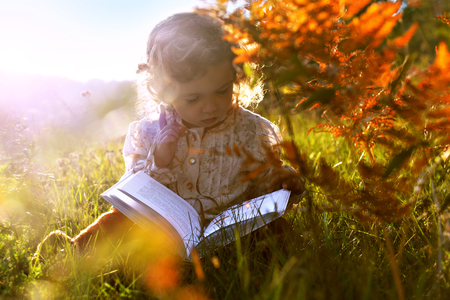 The child in the autumn. A girl with a book is sitting on the grass in the sun at sunset. Portrait of a child in the autumn landscapeの写真素材